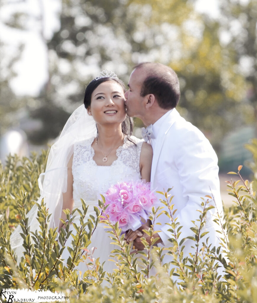 Groom kissing his bride behind tall wildflowers, bouquet in hand